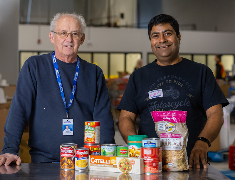 Two Calgary Food Bank volunteers posing with a table of non perishable food such as canned soup, pasta, granola, peanut butter, and canned diced tomatoes.